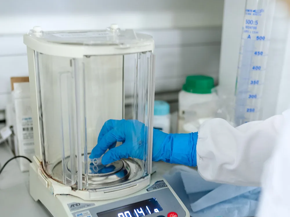 A gloved hand placing a sample on an analytical balance in a laboratory