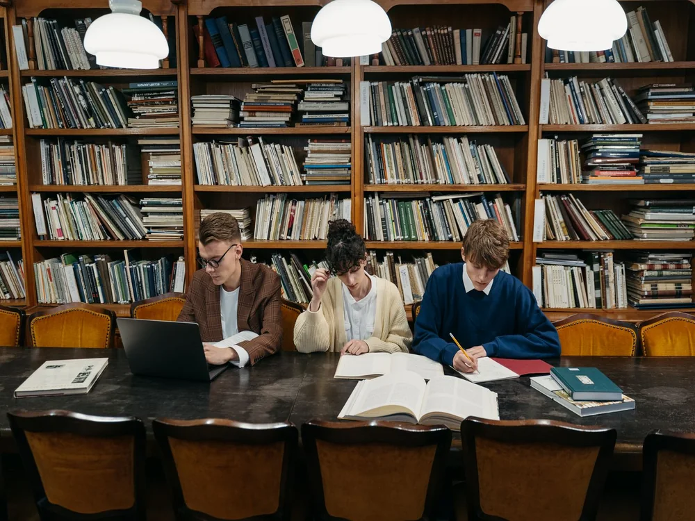 Three students working through books and a laptop at a library table