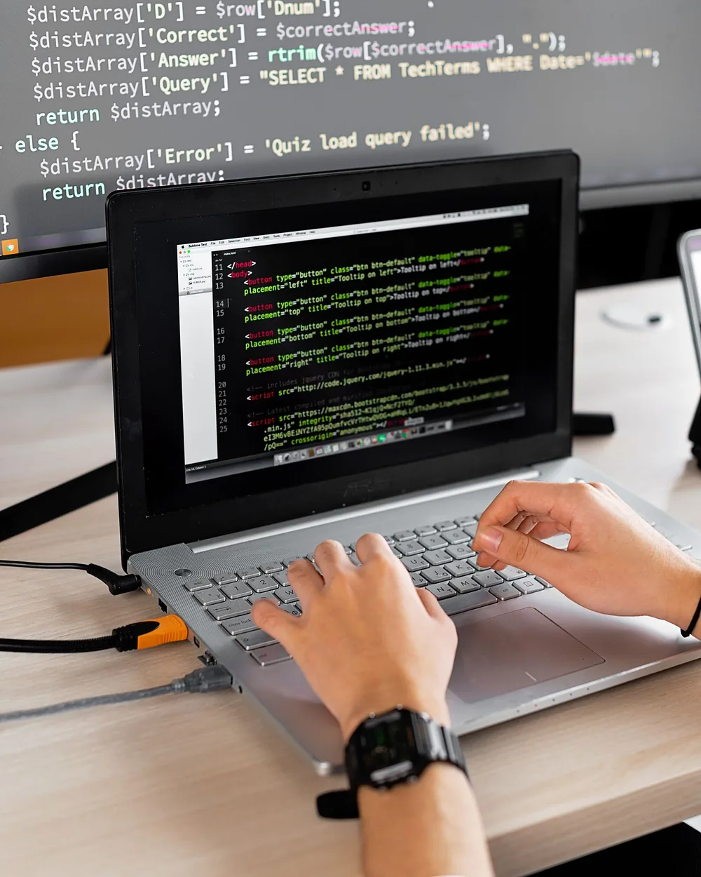 Hands typing code on a laptop, with a monitor full of source code behind it